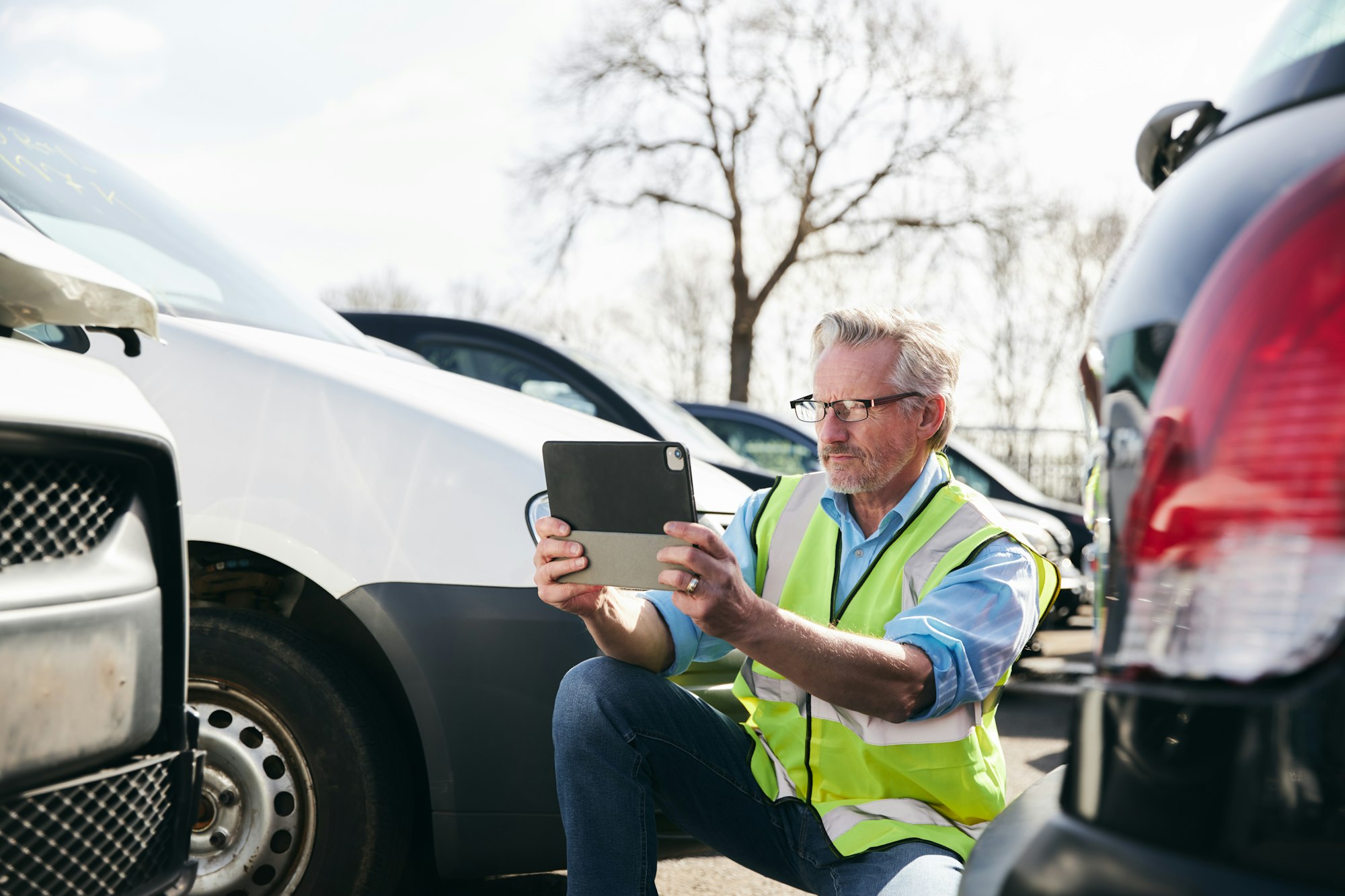 Male Loss Adjuster Taking Photo On Digital Tablet To Assess Insurance Claim In Car Pound