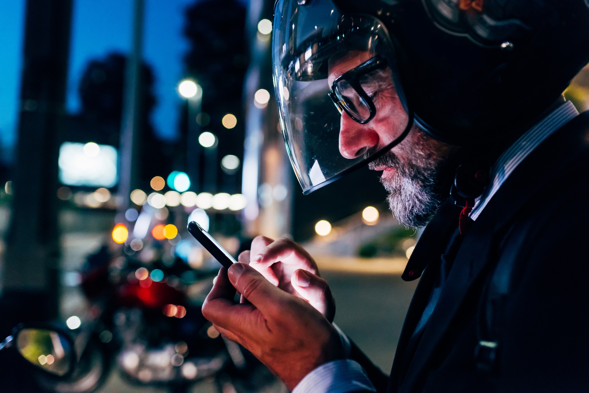 Mature businessman outdoors at night, wearing motorcycle helmet, using smartphone, profile