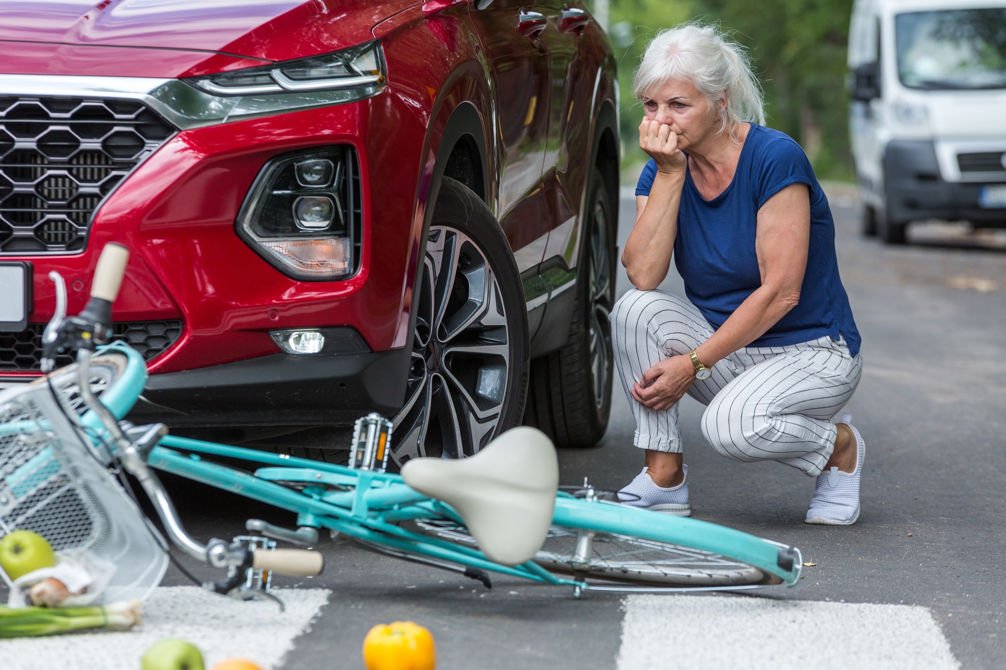 Worried old lady kneels beside her red car after she hits a biker